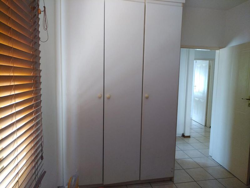 A bedroom featuring built-in cupboards, tiled flooring, and venetian blinds, with a view into the adjacent hallway.
