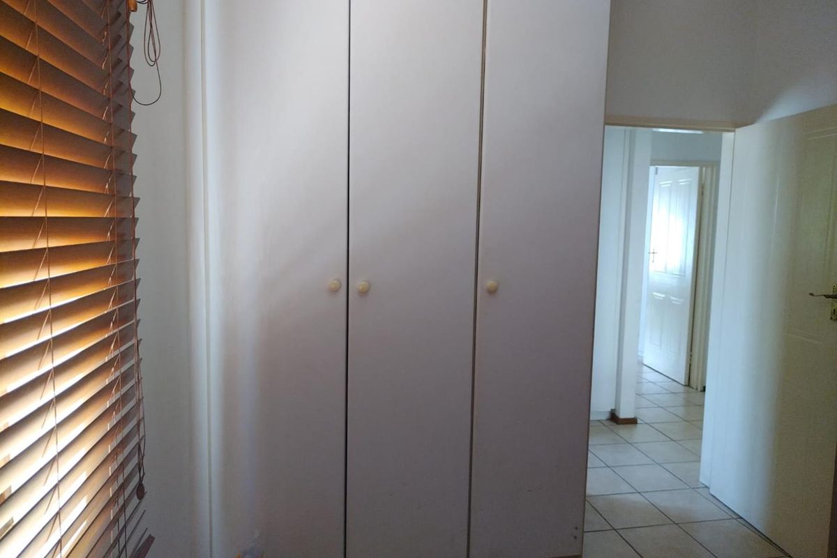 A bedroom featuring built-in cupboards, tiled flooring, and venetian blinds, with a view into the adjacent hallway.