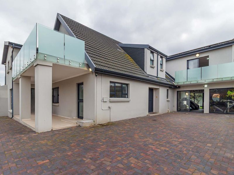 Rear exterior view of the property featuring a paved courtyard, covered patio area, and upper-level balconies with modern glass balustrades.