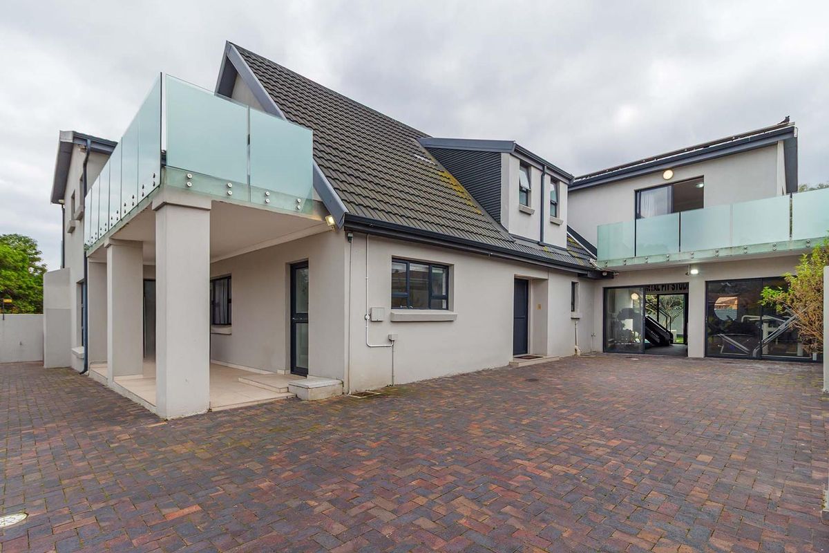 Rear exterior view of the property featuring a paved courtyard, covered patio area, and upper-level balconies with modern glass balustrades.