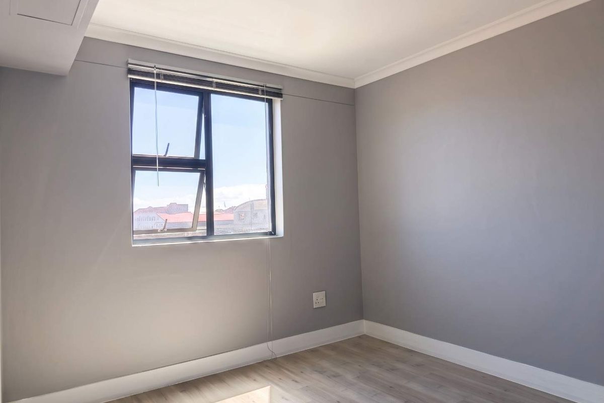 A bright, empty bedroom featuring modern grey walls, laminate flooring, and a large aluminium window with a view.