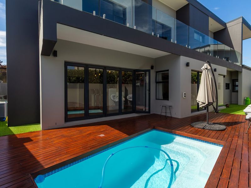 Modern outdoor entertainment area featuring a swimming pool with timber decking, glass balustrades, and seamless access to the interior via bi-fold doors.