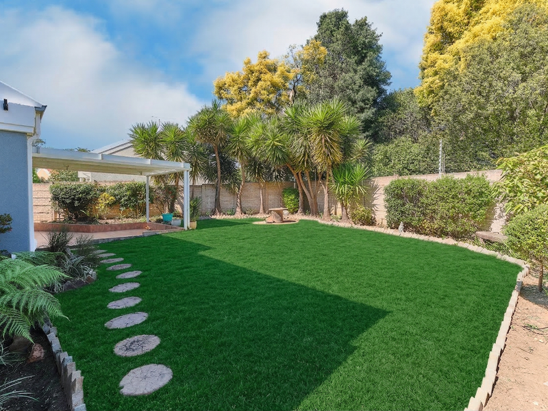 A well-maintained rear garden featuring a lush lawn, established trees, a covered patio area, and a secure boundary wall.