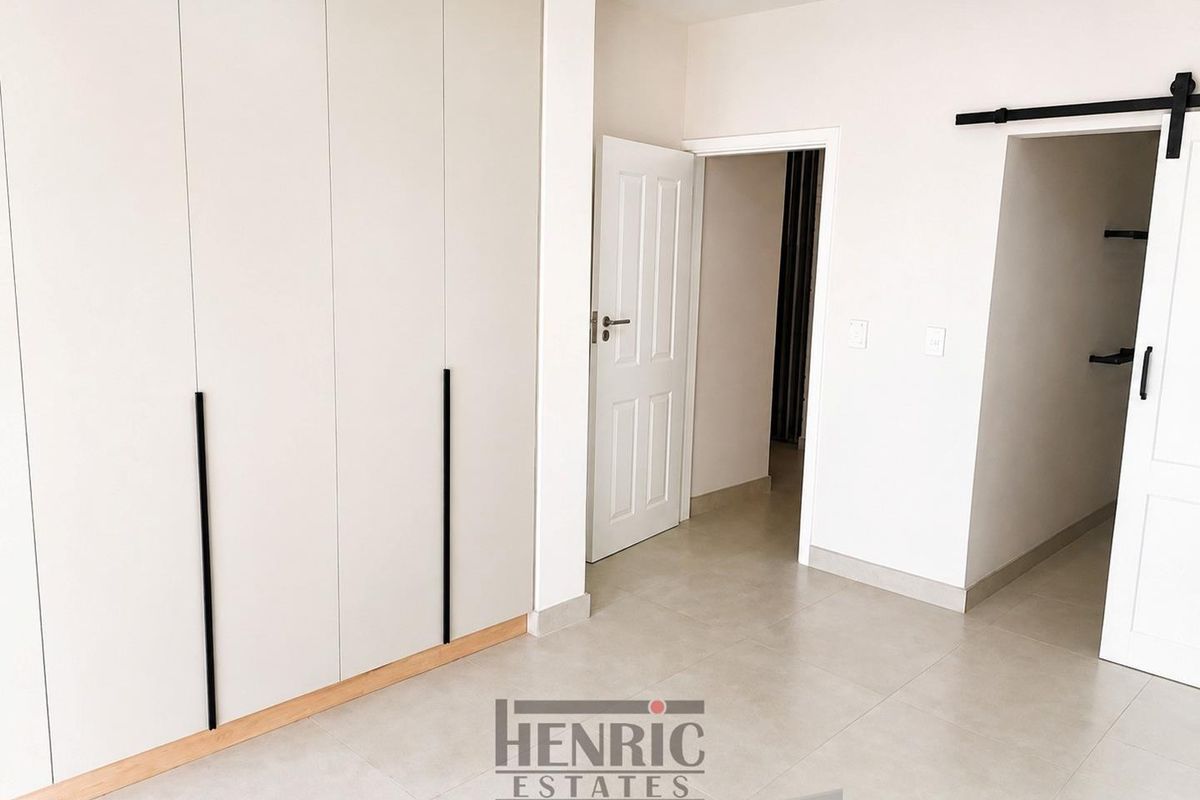 A bright, modern bedroom featuring neutral tiled flooring, floor-to-ceiling built-in cupboards with sleek black handles, and a sliding barn door entrance.
