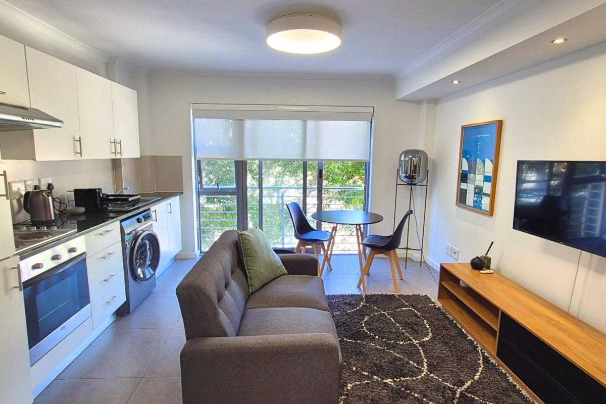Modern open-plan apartment featuring a compact kitchen with built-in appliances, tiled flooring, and a bright living area with direct access to a balcony.