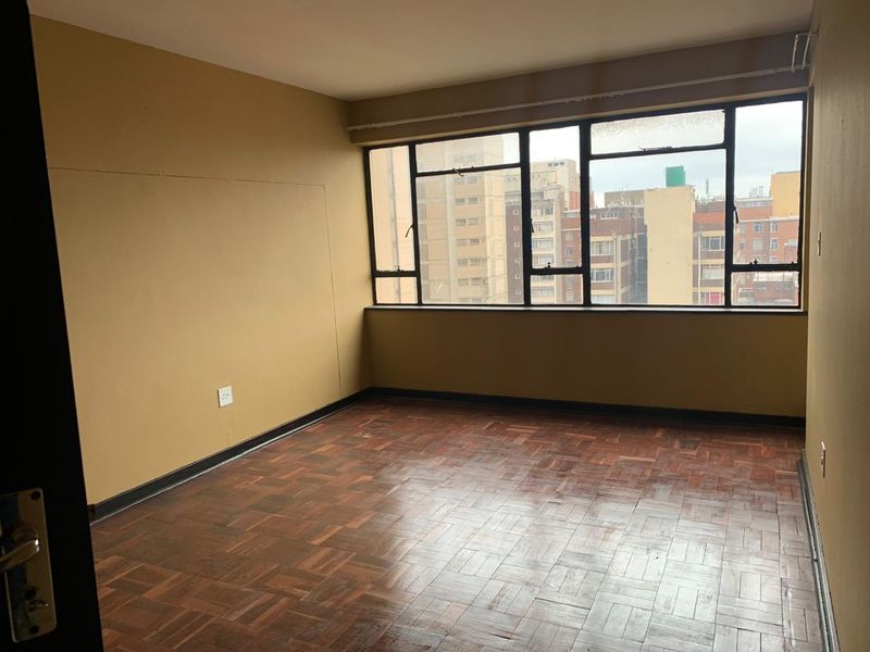 An empty bedroom featuring original parquet flooring, large metal-framed windows with urban views, and neutral wall paint, ready for renovation or staging.