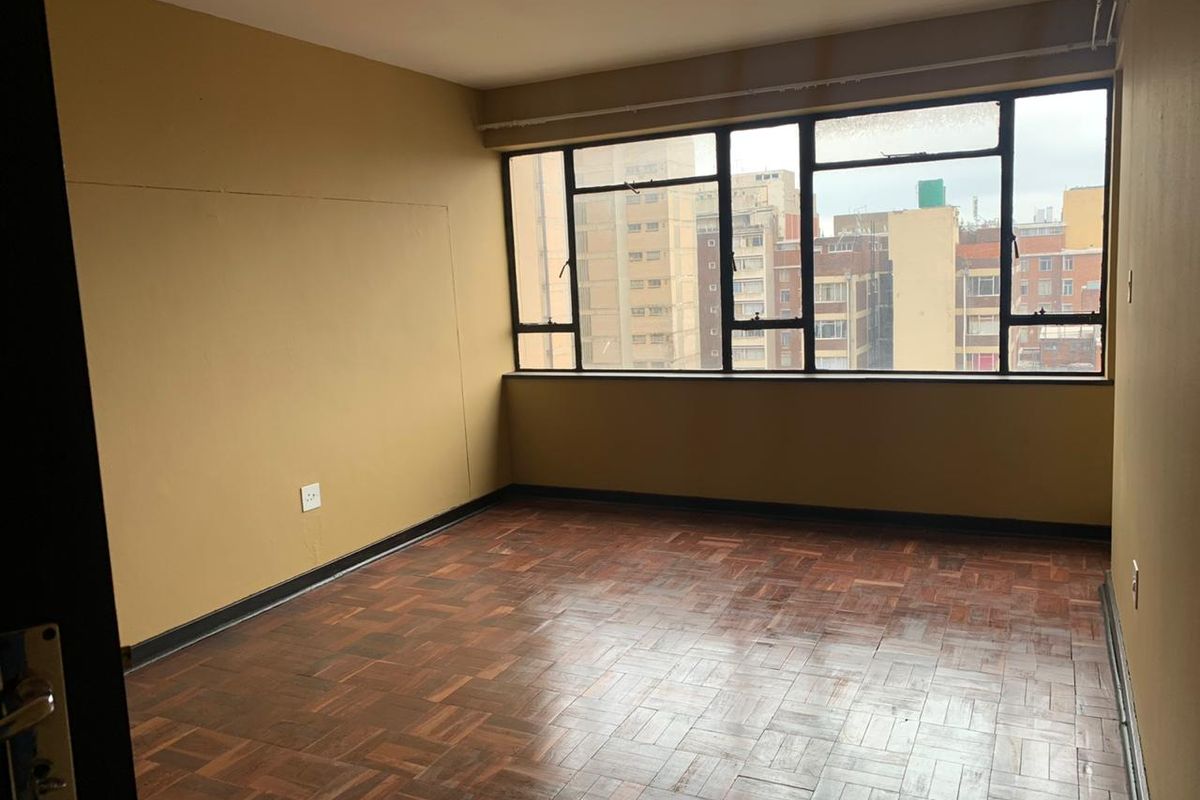 An empty bedroom featuring original parquet flooring, large metal-framed windows with urban views, and neutral wall paint, ready for renovation or staging.