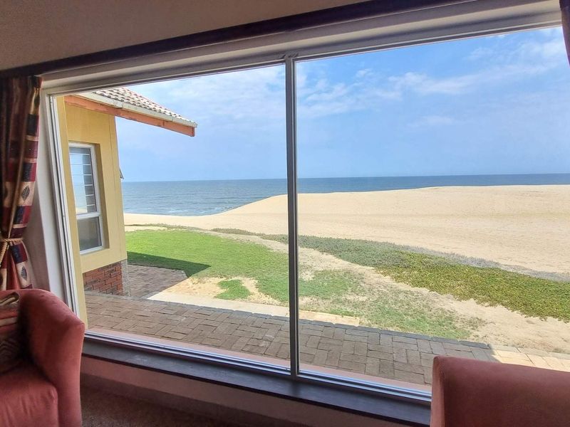 A stunning interior view through a large picture window overlooking a private patio, sandy beach, and the ocean.