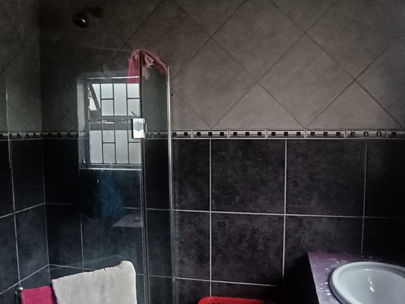 A bathroom featuring a shower enclosure, vanity basin, and dark tiled walls with security bars on the window.