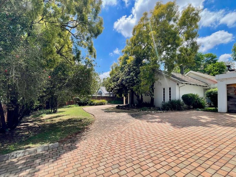 A spacious front exterior view featuring a large paved driveway, mature trees, and a well-maintained garden leading to the main house entrance.