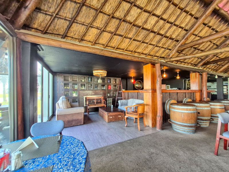 A rustic bar area featuring a traditional thatched roof, exposed timber support beams, built-in bar counter, and extensive shelving, creating a warm, atmospheric space.