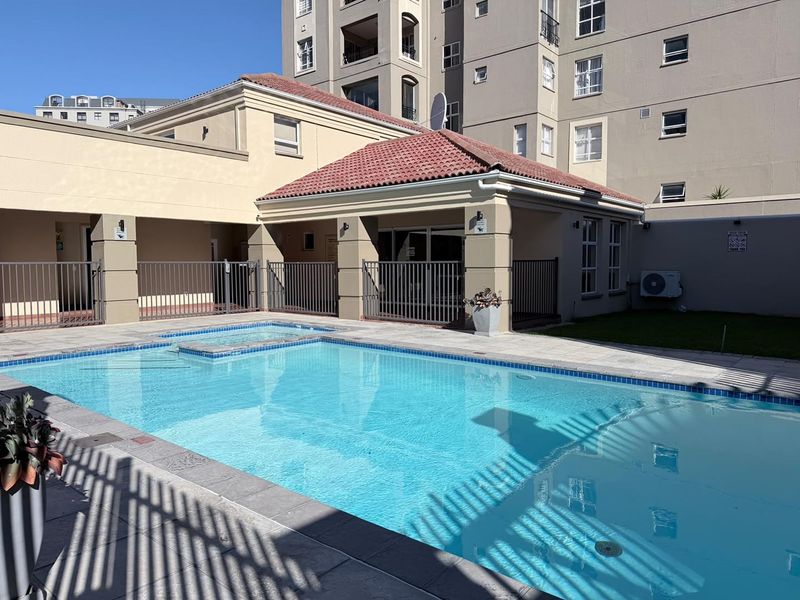 A well-maintained communal swimming pool area featuring paved surrounds, metal safety fencing, and a covered patio structure within an apartment complex.