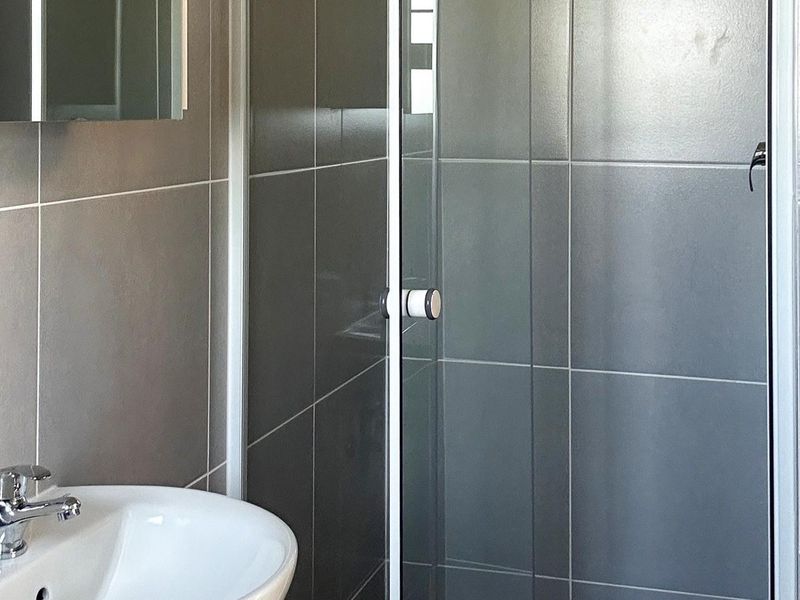 A clean, modern bathroom featuring grey wall tiles, a glass shower enclosure, and a white pedestal basin with chrome fixtures.