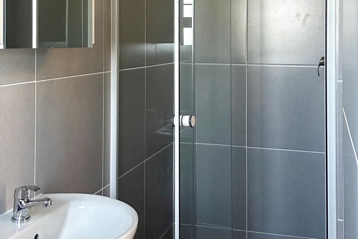 A clean, modern bathroom featuring grey wall tiles, a glass shower enclosure, and a white pedestal basin with chrome fixtures.