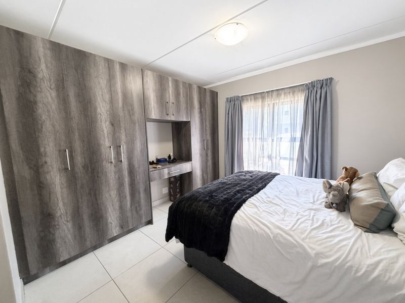 A well-maintained bedroom featuring extensive built-in cupboards with an integrated vanity unit, neutral tiled flooring, and large windows with curtains.