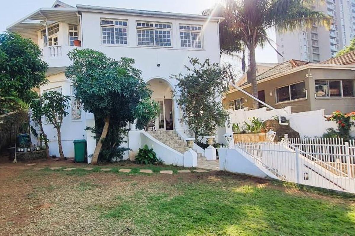 A view of the rear exterior of a multi-story house featuring a white facade, arched entrance, and a garden area with a white perimeter fence.