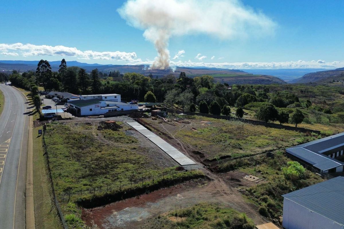 Aerial view of a commercial vacant land plot featuring a concrete driveway, perimeter fencing, and adjacent commercial buildings with surrounding landscape.