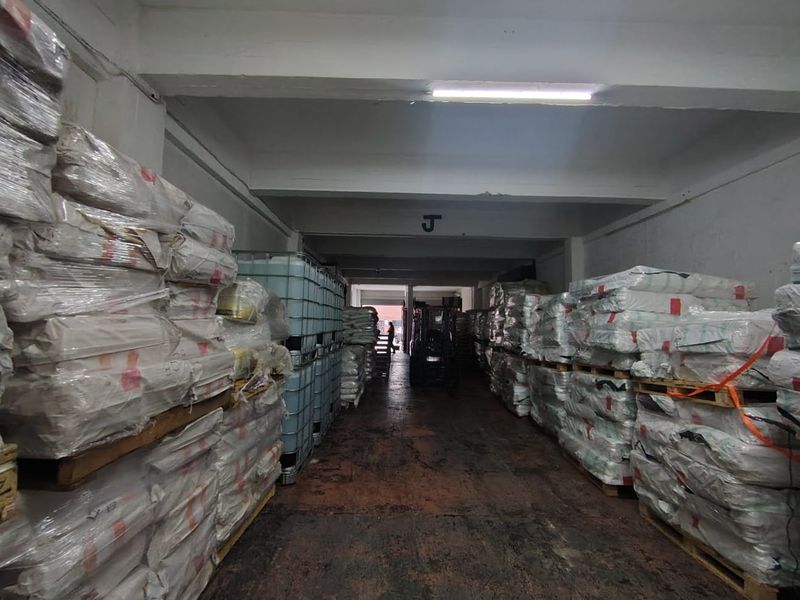 Industrial warehouse storage area featuring concrete flooring, exposed structural beams, and overhead fluorescent lighting, currently utilized for palletized goods storage.