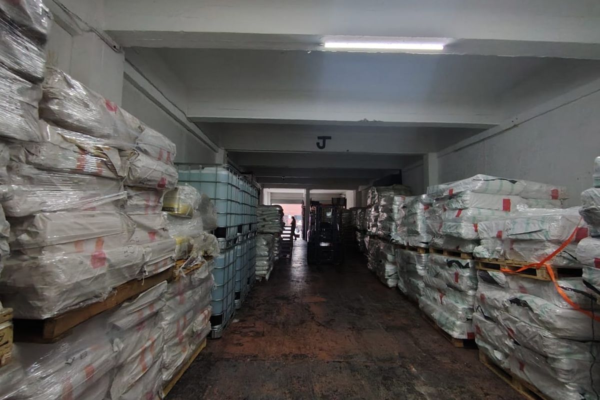 Industrial warehouse storage area featuring concrete flooring, exposed structural beams, and overhead fluorescent lighting, currently utilized for palletized goods storage.