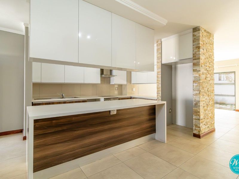 Modern kitchen featuring high-gloss white cabinetry, a wood-accented breakfast bar, stone cladding feature wall, and neutral tiled flooring in an open-plan layout.