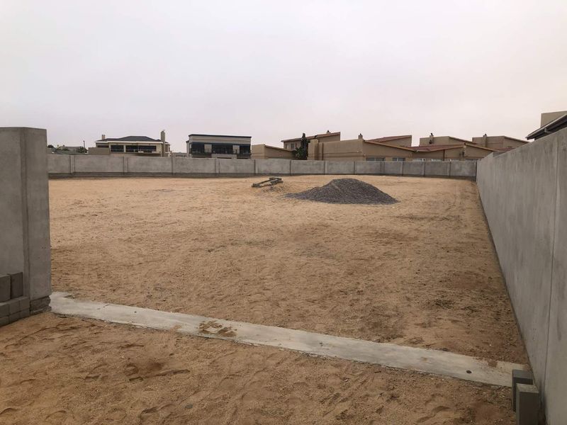 A vacant residential plot enclosed by precast concrete boundary walls, featuring a level sandy surface and a concrete foundation strip.