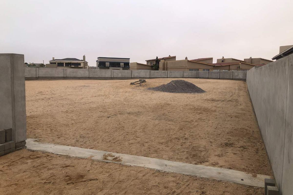 A vacant residential plot enclosed by precast concrete boundary walls, featuring a level sandy surface and a concrete foundation strip.