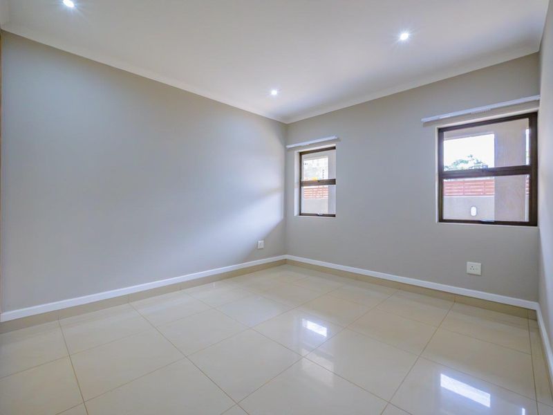 A bright, empty bedroom featuring neutral wall tones, polished tiled flooring, recessed downlights, and built-in cupboards with aluminium-framed windows.