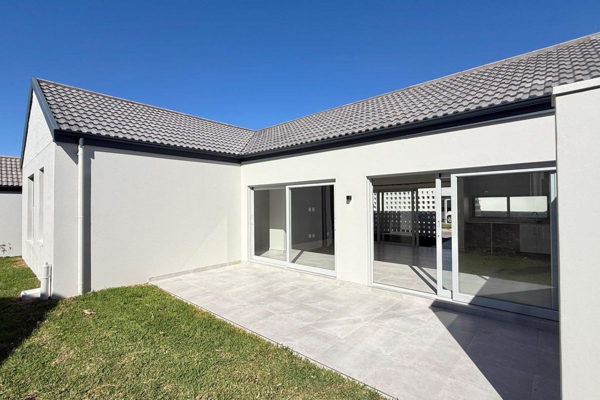 A modern rear exterior view featuring a tiled patio, large sliding glass doors, and a well-maintained lawn under a clear blue sky.