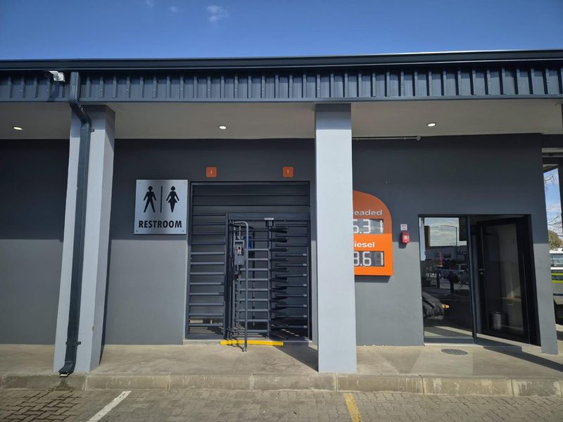 Exterior view of a commercial fuel station facility featuring a secure turnstile entrance to restrooms and a digital fuel price display board.