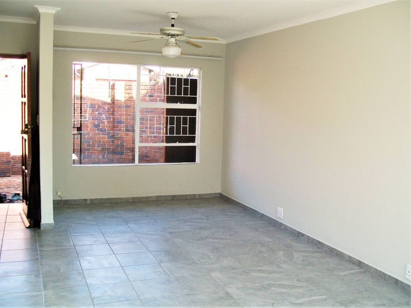 A bright, empty living area featuring neutral walls, tiled flooring, and a ceiling fan, with a window overlooking an exterior brick wall.