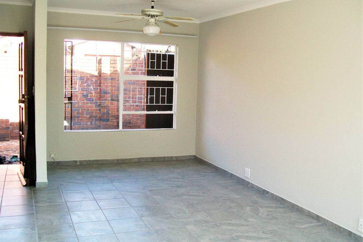 A bright, empty living area featuring neutral walls, tiled flooring, and a ceiling fan, with a window overlooking an exterior brick wall.