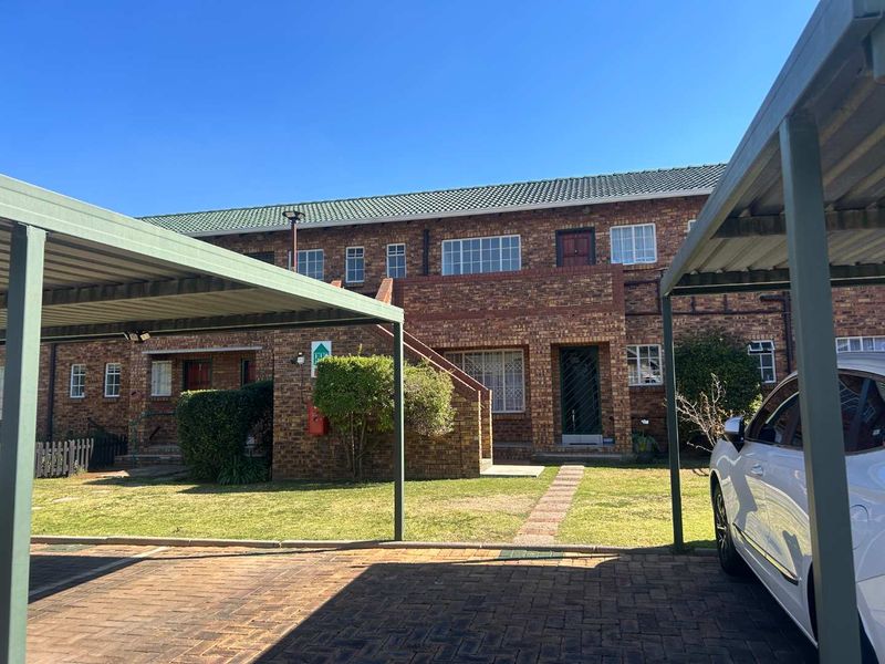 Front view of a brick townhouse complex featuring covered carports, paved driveway, and established garden area.