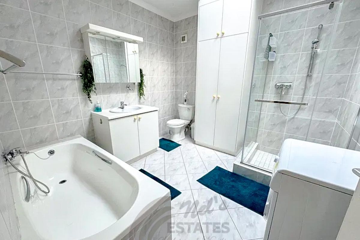 A well-appointed bathroom featuring a built-in bathtub, separate glass-enclosed shower, vanity cabinet with basin, toilet, and tall storage cabinetry with tiled finishes.
