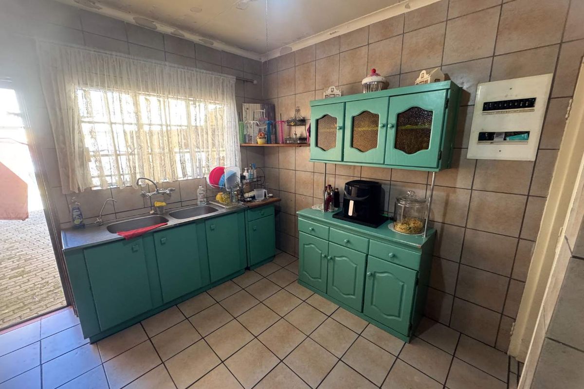 A dated kitchen featuring teal painted cabinetry, double stainless steel sink, tiled walls and flooring, and an integrated electrical distribution board.