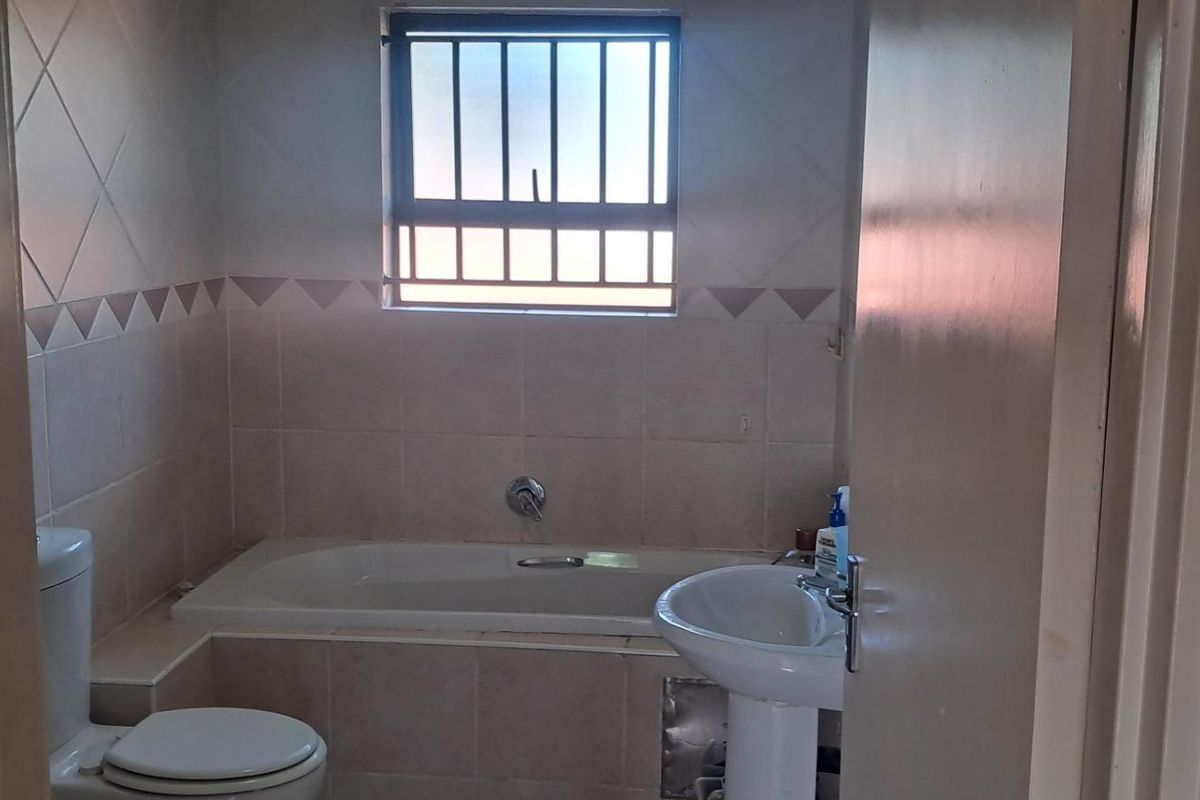 A functional bathroom featuring a built-in bathtub, pedestal basin, and toilet, finished with neutral wall tiling and a security-barred window.