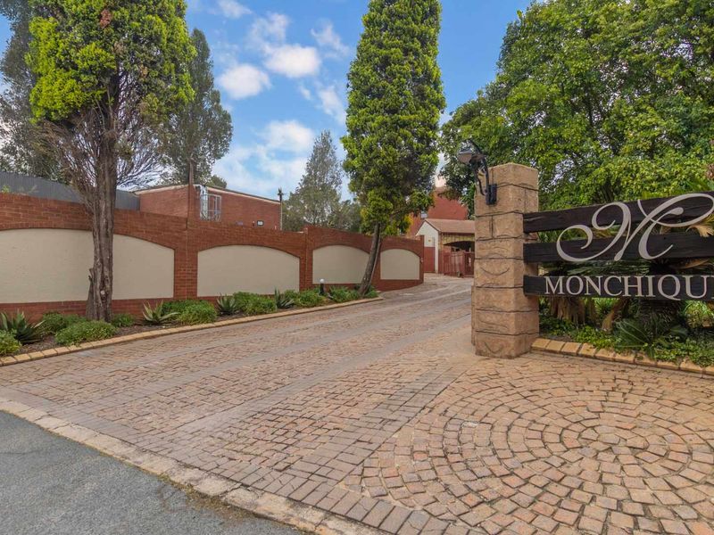 The entrance to Monchique complex featuring a paved cobblestone driveway, stone pillars, and a brick boundary wall with established landscaping.