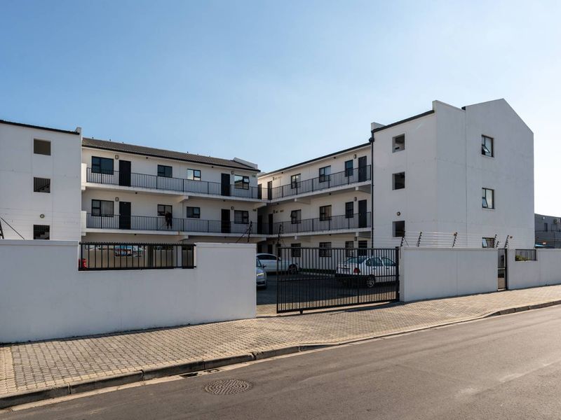 Exterior view of a multi-story apartment complex featuring white facade, paved driveway, secure perimeter wall with electric fencing, and balcony access.