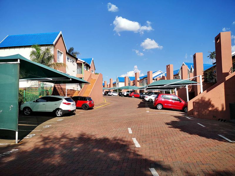 Exterior view of a townhouse complex featuring a paved driveway, designated parking bays with shade-cloth carports, and multi-level residential units.