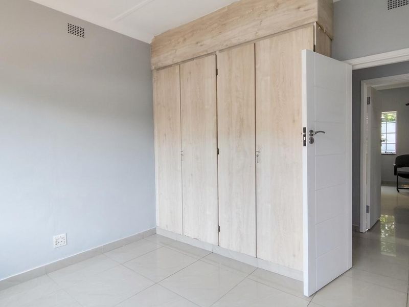 A bright, empty bedroom featuring light wood-finish built-in cupboards, neutral grey walls, and clean tiled flooring.