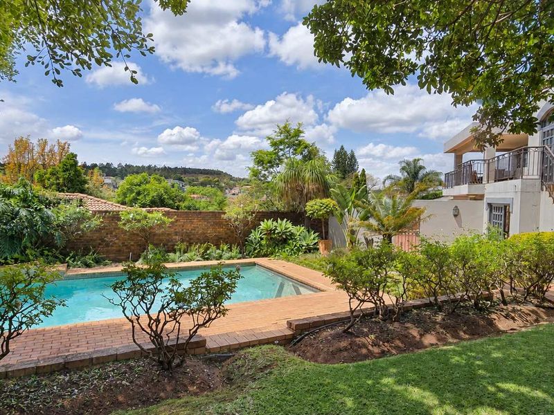 A well-maintained outdoor pool area featuring brick paving, lush garden landscaping, and an external staircase leading to an upper-level balcony.