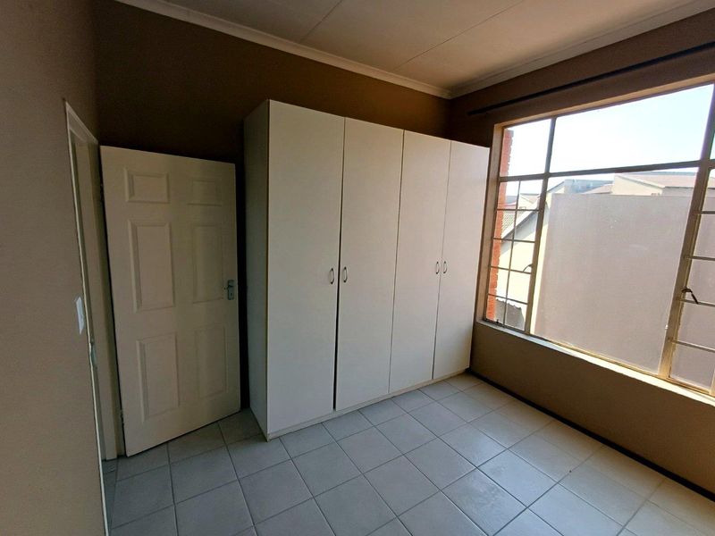 A bright, empty bedroom featuring neutral tiled flooring, built-in white cupboards, and a large window providing natural light.