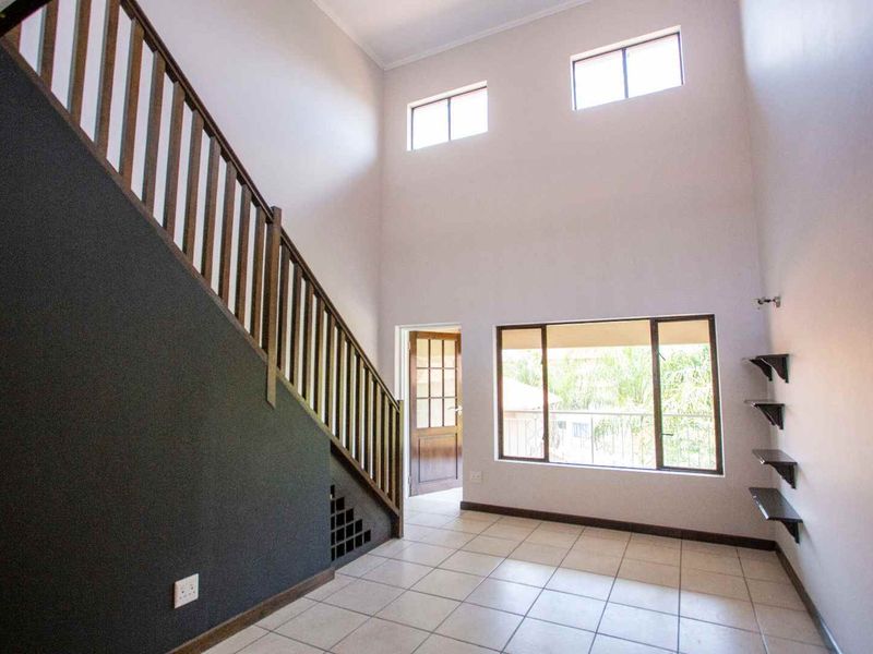 Spacious double-volume living area featuring tiled flooring, a wooden staircase with balustrade, and large windows providing ample natural light.