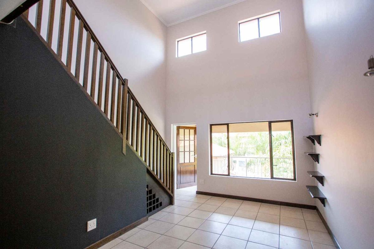 Spacious double-volume living area featuring tiled flooring, a wooden staircase with balustrade, and large windows providing ample natural light.