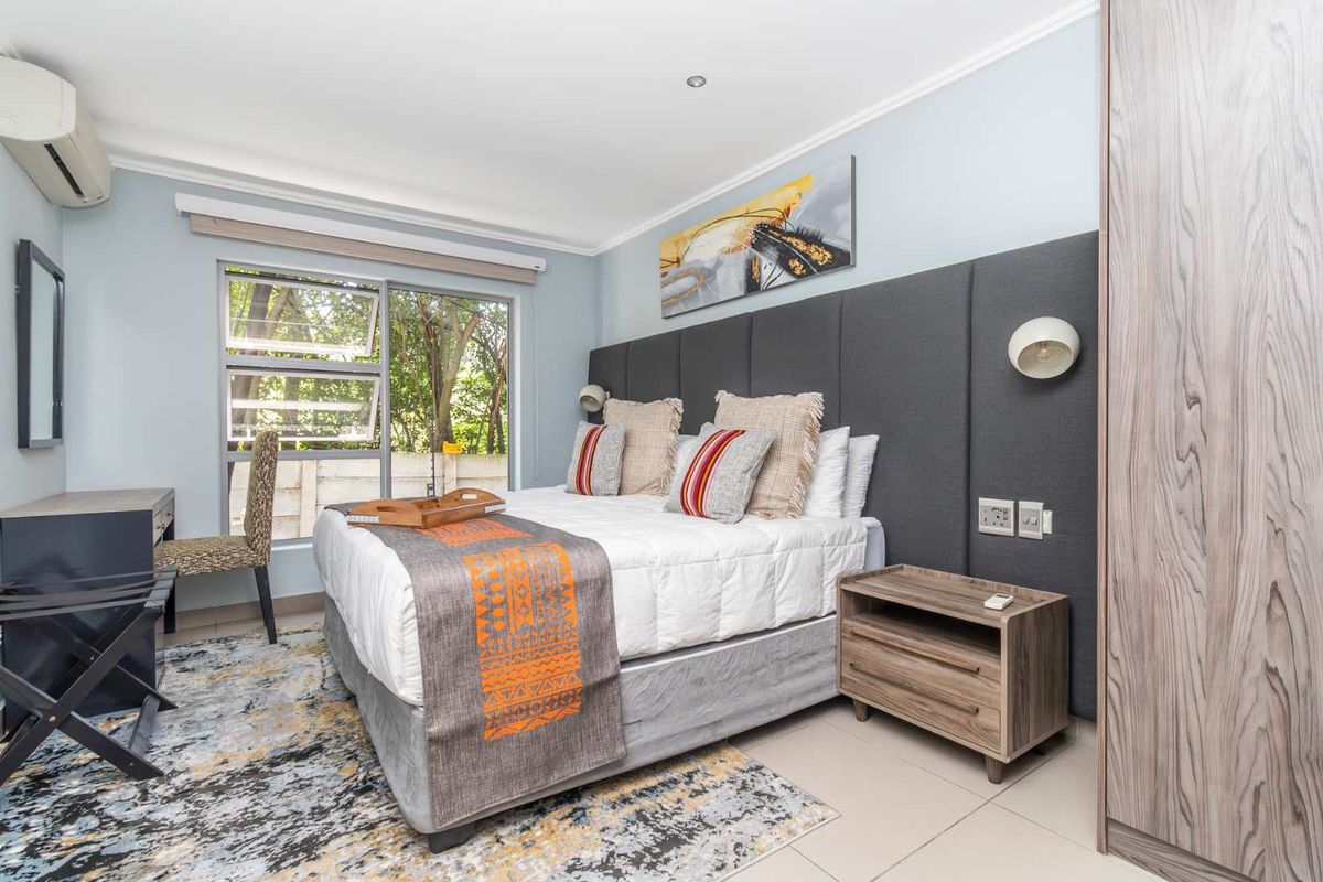 A well-maintained bedroom featuring a large upholstered headboard, built-in wardrobe, tiled flooring, and a wall-mounted air conditioning unit.
