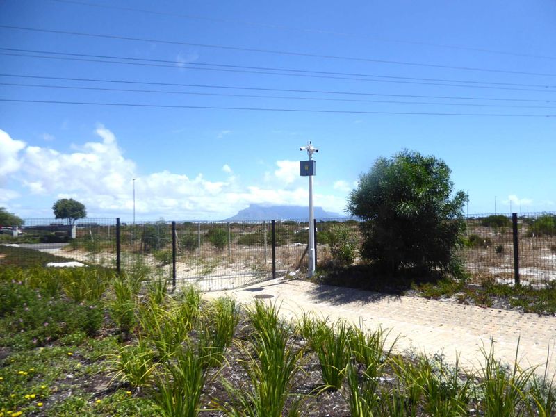 A view of a vacant residential plot featuring a paved entrance, security camera, and wire mesh perimeter fencing with distant mountain views.