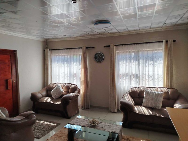 A living room featuring tiled flooring, a drop ceiling, and large windows with security gates, providing a functional and well-lit space.