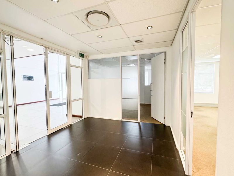 A bright, vacant office space featuring modern glass partitions, suspended ceilings with recessed lighting, and clean tiled flooring, suitable for a professional business environment.