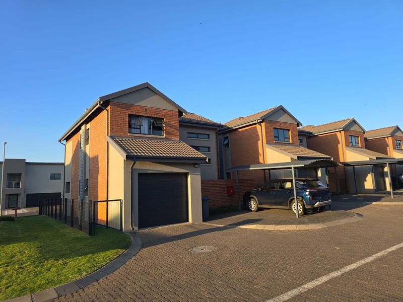 Front elevation of a modern townhouse featuring a brick and plaster facade, single garage, carport, and paved driveway within a residential complex.