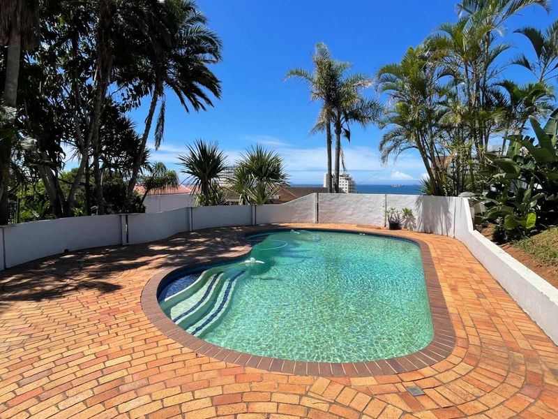 Outdoor swimming pool area featuring brick paving, perimeter boundary walls, and established tropical landscaping with a distant ocean view.