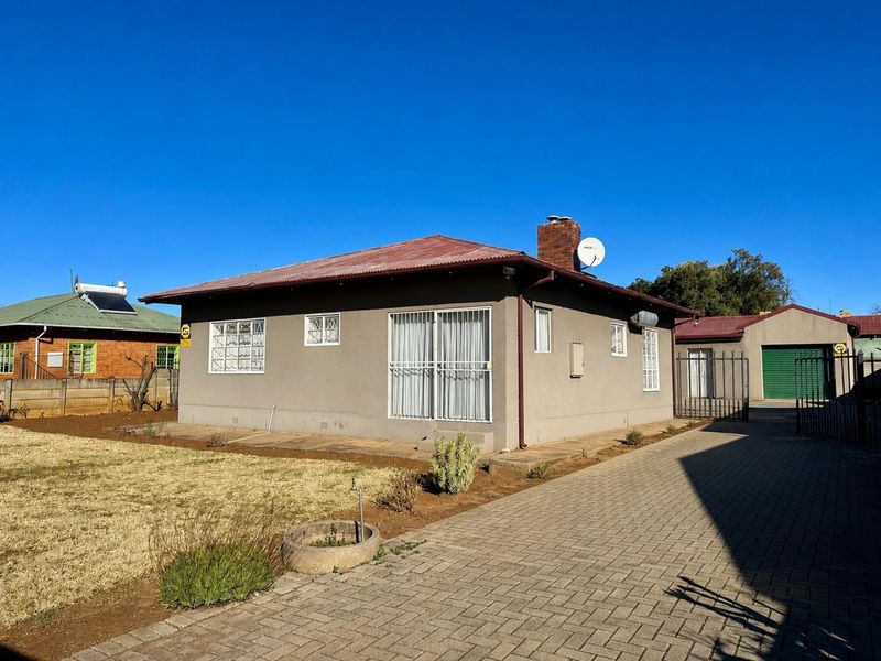 Rear view of a single-story house featuring a corrugated metal roof, brick chimney, paved driveway, and security features including window bars and gates.